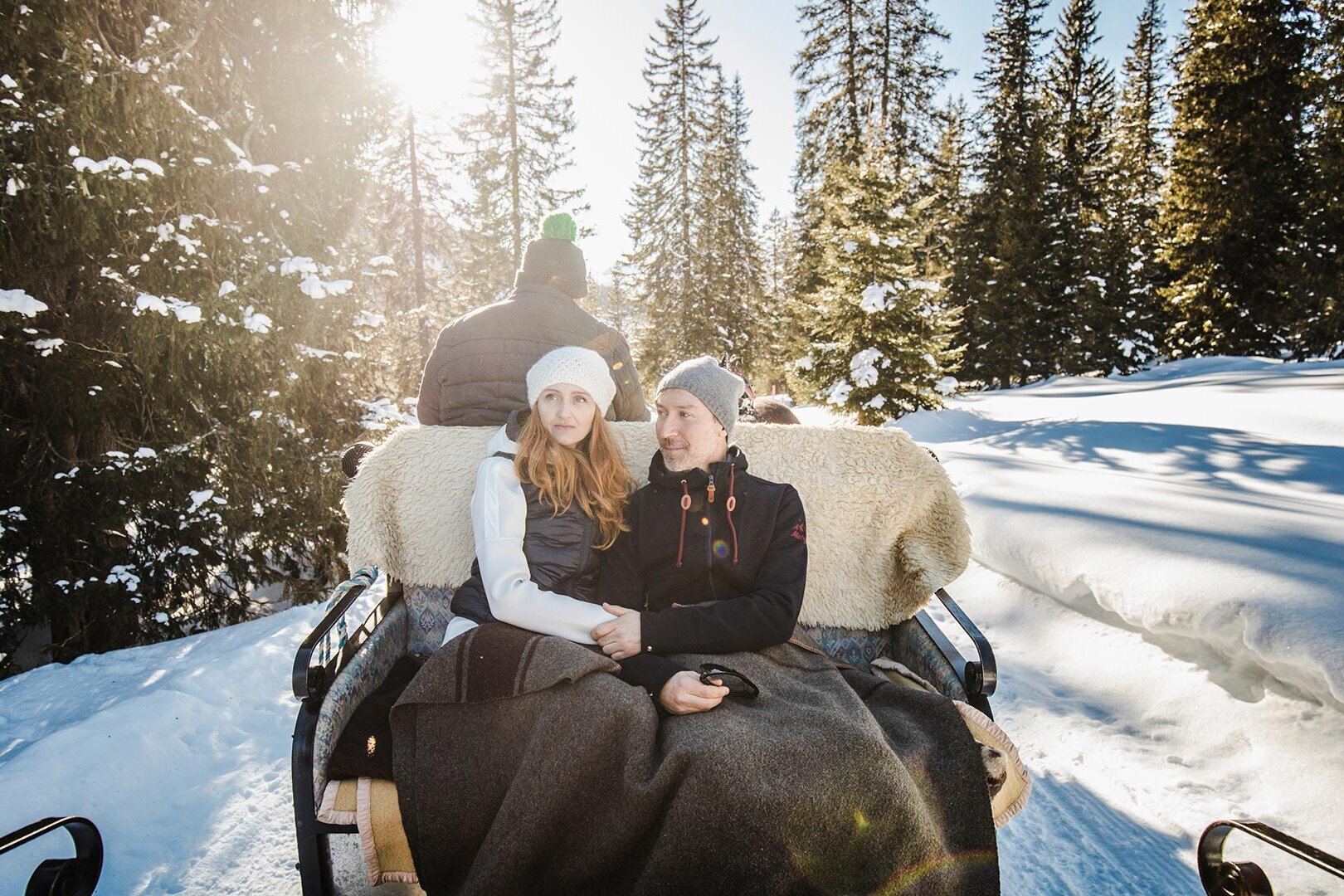Zwei Frauen sitzen warm eingehüllt auf einer Pferdeschlittenfahrt durch verschneiten Winterwald bei Sonnenlicht.
